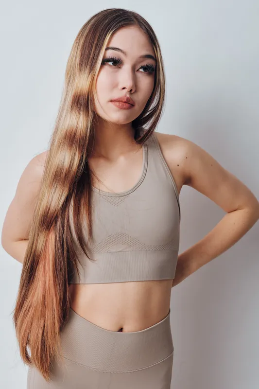 Young woman with long brown hair wearing beige sportswear posing with arms behind back against white background for fitness and wellness concept.