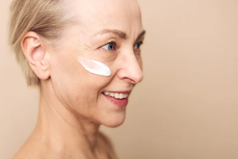 Mature woman with short hair applying cream on her cheek, smiling with clear skin. Concept of skincare, anti aging, beauty routine, wellness, and natural care.