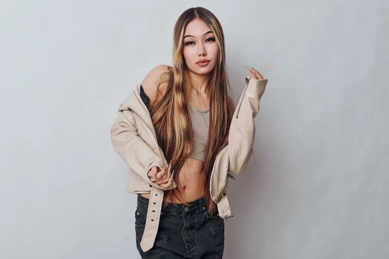 Young woman in casual outfit posing confidently with beige jacket and crop top against plain background. Fashion model with long hair and relaxed expression.