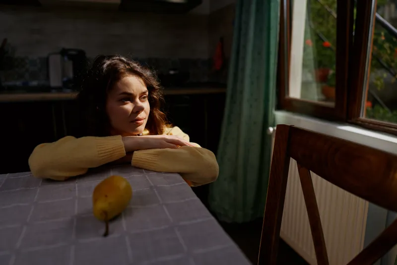 Young woman rests her chin on folded arms at a table by a window, pear on the table, warm natural light shaping a calm indoors mood and everyday lifestyle moment.