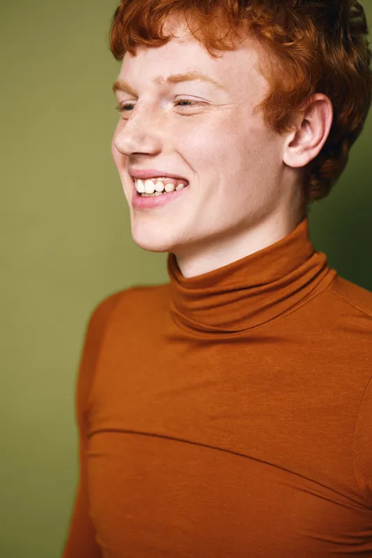 Young man with red hair wearing brown turtleneck smiling and looking to side, portrait of happy person with freckles and casual style on green background.
