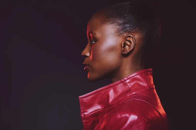 Profile portrait of a young black woman with dark skin wearing a red leather jacket. Studio shot with dramatic lighting on black background. Concept of fashion and beauty.