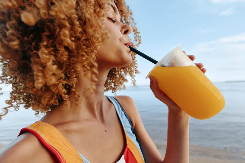A young woman with curly hair drinks orange juice through a straw on a sunny beach, water glistening in the background, conveying a moment of relaxed summer refreshment.