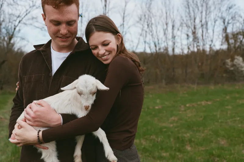 A couple outdoors gently holds a young goat kid in a rural field, sharing a loving embrace amid meadow and trees, a warm lifestyle moment full of affection and happiness