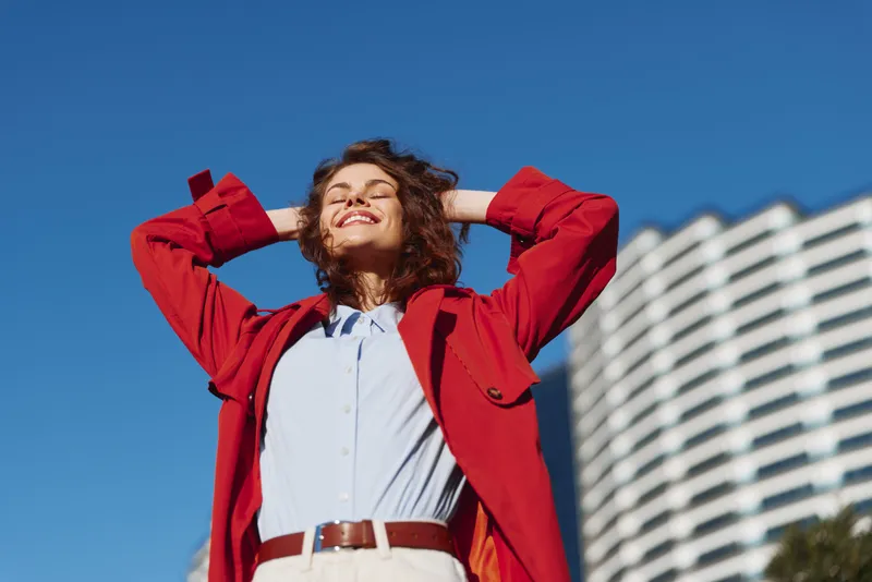 Cheerful woman outdoors, wearing a red jacket and white shirt, basking in sunlight with a relaxed pose against a clear blue sky and urban backdrop