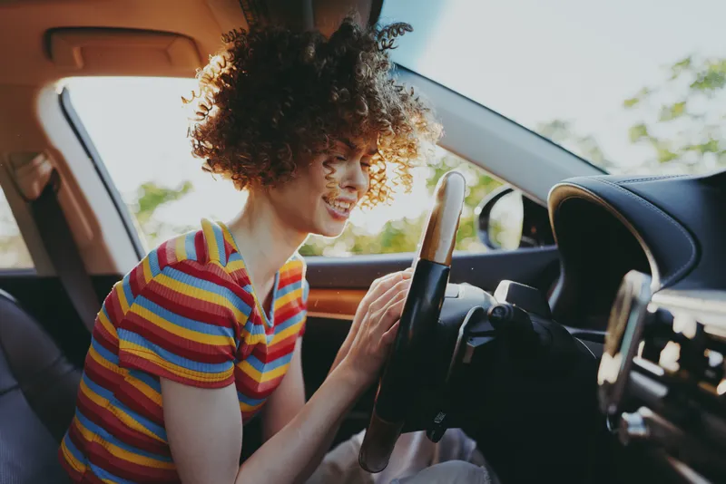 Smiling woman driving a car with a striped shirt, inside a sunny vehicle. Focus on lifestyle, casual road trip, everyday wellness, retro color tones create a warm, nostalgic mood.