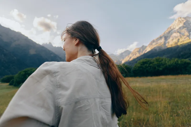 A woman with long hair tied in a ponytail stands outdoors in a grassy field surrounded by mountains under a partly cloudy sky, enjoying a peaceful lifestyle moment.