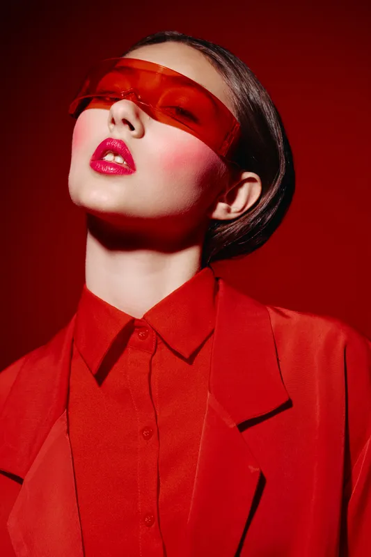 Close-up of a stylish woman wearing red tinted visor and red jacket. Bold makeup with vibrant lip color and blush stands out against deep red background. Concept of fashion creative beauty.
