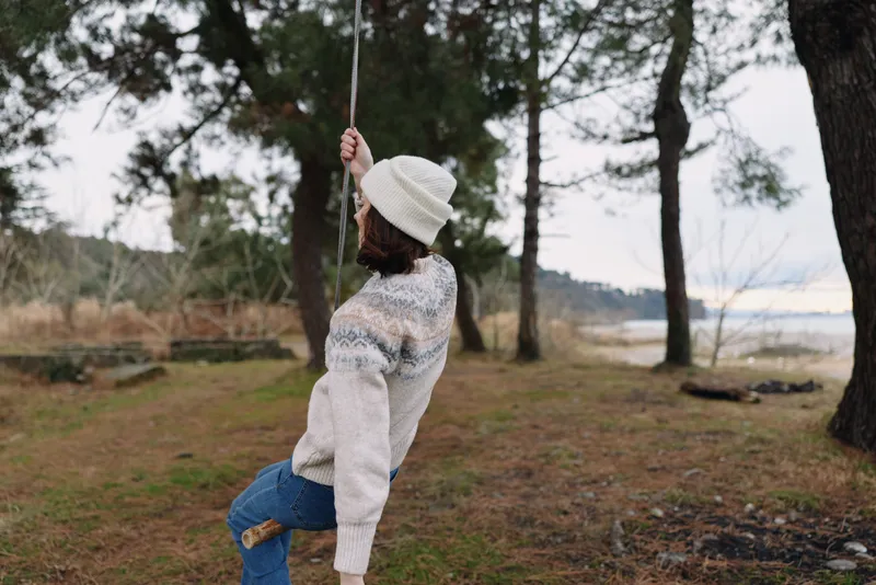 Outdoor scene with a person on a rope swing wearing a knitted sweater and beanie, in a forested lakeside setting. A calm, cozy moment of outdoor winter leisure.