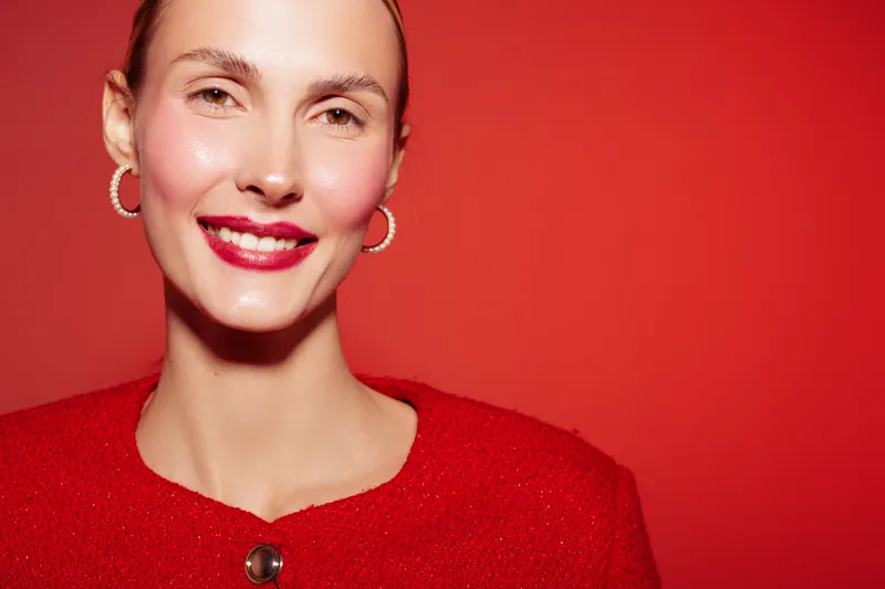 Close-up portrait of smiling woman with red lipstick and earrings wearing red sweater on red background. Concept of beauty, fashion, confidence, and vibrant style.