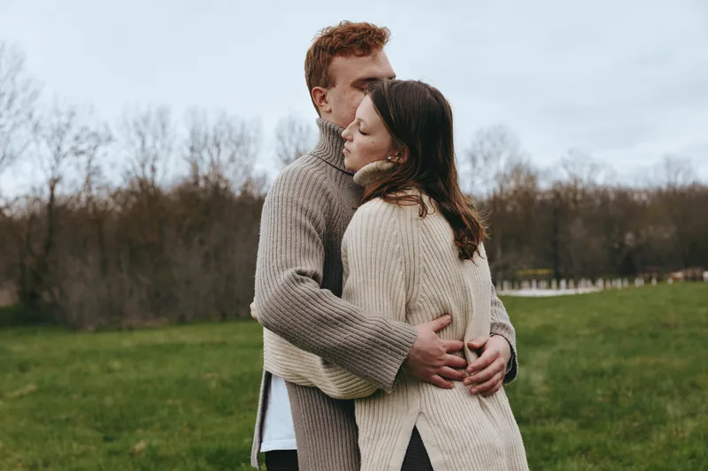 A couple embraces in a grassy park, wearing cozy knits, capturing intimate connection and warmth in a natural outdoor scene that highlights togetherness, winter charm, and lifestyle storytelling.