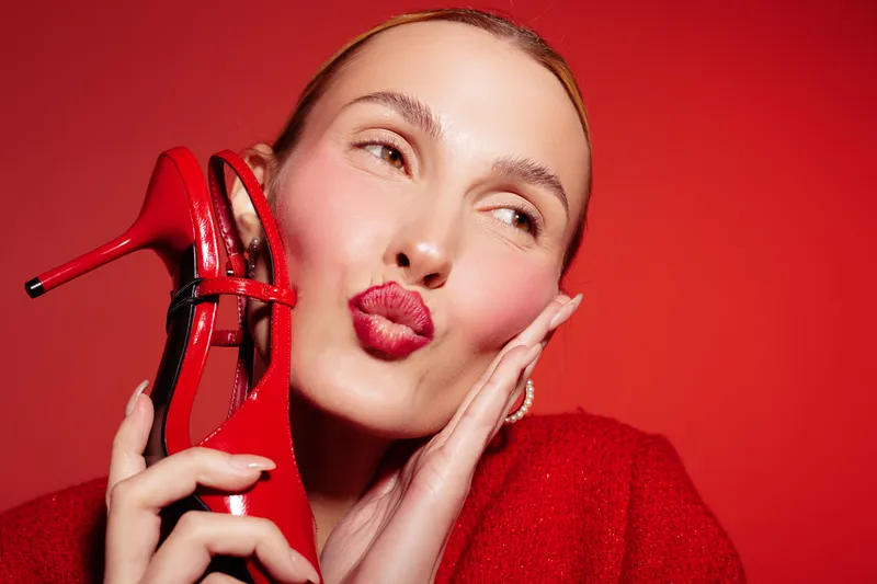 Young woman with red lipstick holds red high heel shoe near face, posing on red background. Concept of fashion, beauty, glamour, and style with vibrant color contrast.