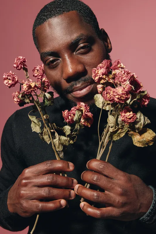 African man holding dried roses with closed eyes in creative conceptual visual style, portrait with dark skin and emotional expression on pink background for artistic photography.