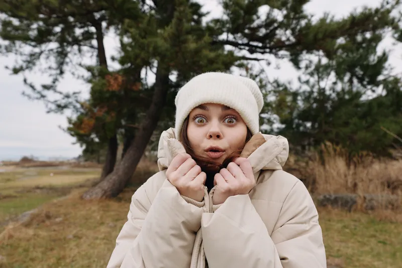 Woman in a beige winter coat and beanie outdoors, surprised expression as she pulls the collar tight in a chilly park scene, natural light