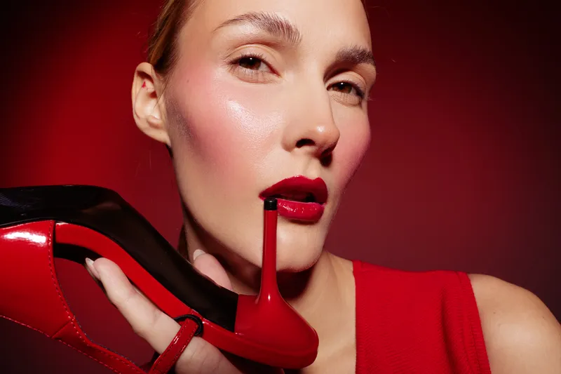 Close-up of woman with glossy red lips holding red high heel shoe near face on red background. Concept of fashion, beauty, glamour, and bold style with vibrant colors and confident mood.