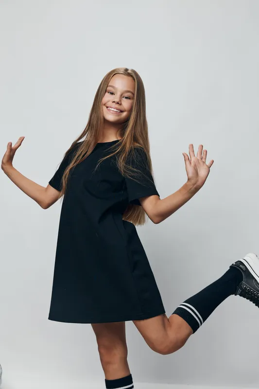 Girl smiling and posing with one leg bent wearing black dress and sneakers against a light background. Youthful female with long hair in casual sporty outfit standing joyful.