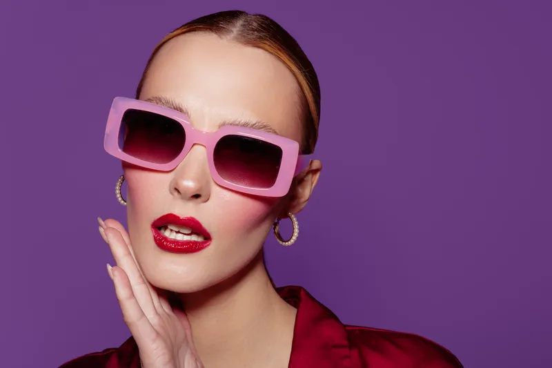 Stylish woman with pink sunglasses and red lipstick poses on purple background. Fashion portrait with glossy makeup and elegant earrings. Concept of beauty and glamour.