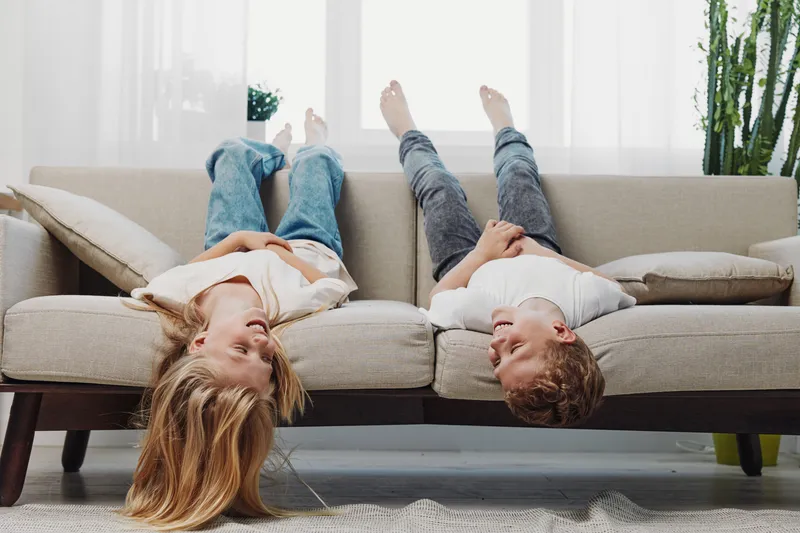 Two children lie upside down on a beige sofa in a bright living room. The playful siblings enjoy a relaxed moment together, creating a joyful and carefree atmosphere at home.