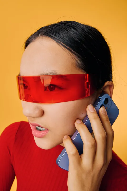 Woman with braces talking on smartphone wearing futuristic red visor and red top on yellow background closeup. Technology communication emotion and modern style with focus on face and hand.