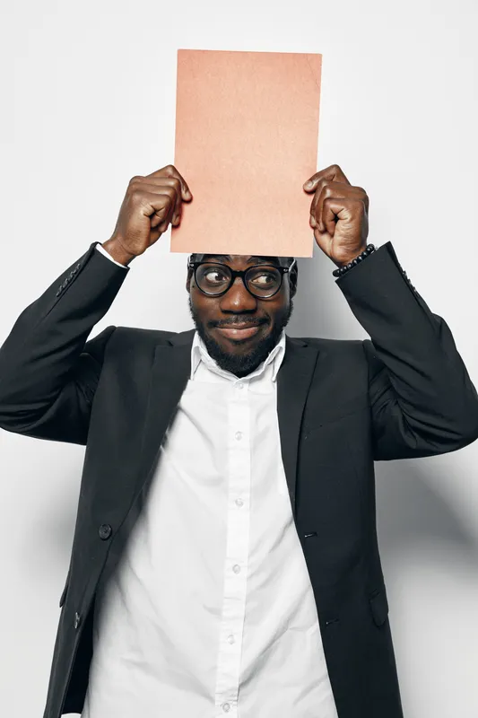 African man with dark skin and glasses holding blank paper above head in black jacket and white shirt. Studio portrait with confident expression on white background.