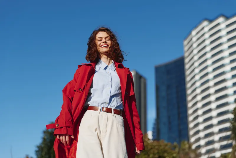 Smiling young woman with curly hair wearing a red coat and light blue shirt stands in a sunny urban street, posing confidently as bright city energy surrounds her.