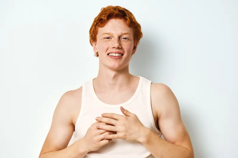 Young man with red hair smiling and clasping hands on chest wearing white tank top against white background. Portrait, happy expression and casual clothing in studio setting.
