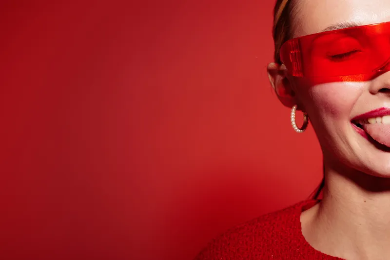 Close-up of smiling woman wearing red sunglasses and red sweater against red background. Playful expression with tongue out. Concept of fun, fashion, and vibrant style.