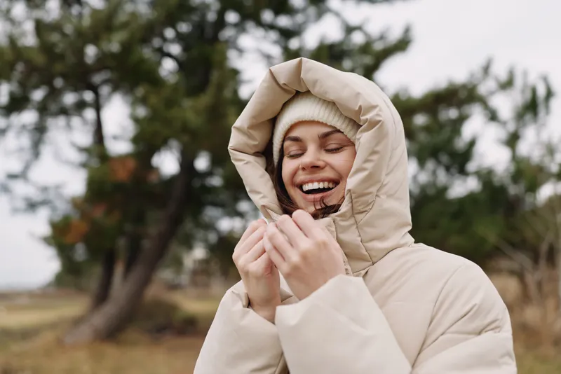 Woman in a beige winter coat and hood smiling warmly outdoors on a cold day, enjoying nature, fresh air, and subtle sunlight.