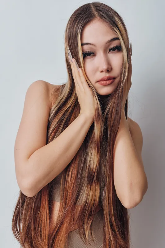 Woman with long straight hair touching face with hands, posing in front of light background for beauty, fashion and hairstyle concept portrait indoors.