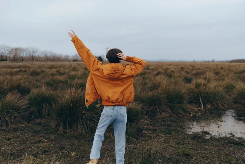 Woman outdoors in countryside field wears jacket and jeans, lifestyle spring moment facing the sky, calm walking pose, overcast light, windy rural nature background, wide horizon