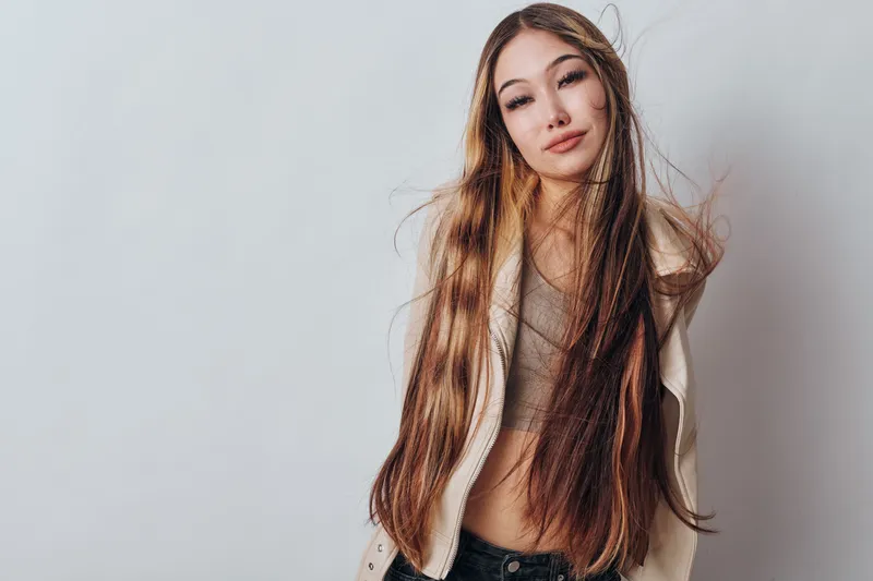 Young woman with long straight hair wearing light jacket and casual top stands against white background with relaxed expression and closed eyes for fashion portrait.