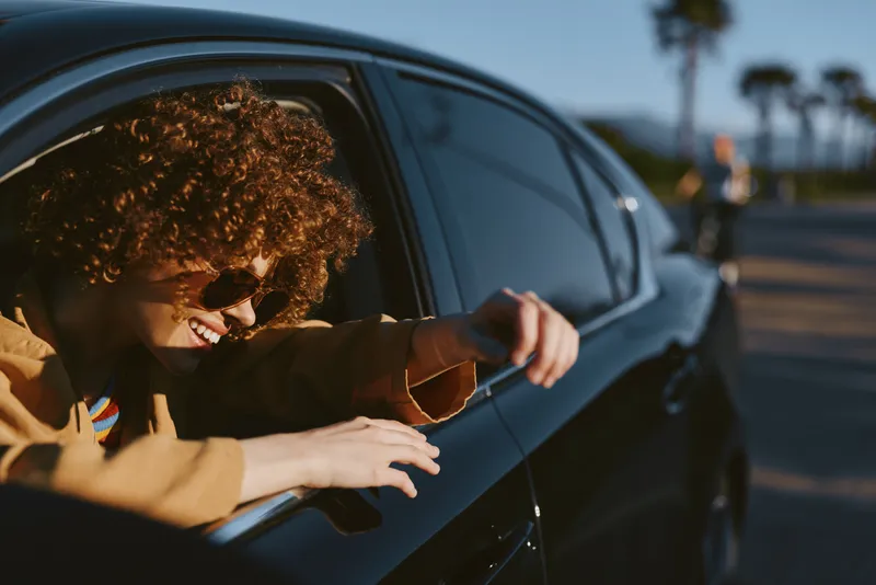 Joyful woman with curly hair leans from a moving car window, wearing sunglasses, on a carefree urban drive, captured in warm retro film color and bright daylight.