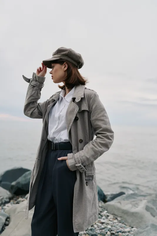 Young woman in trench coat and hat stands on rocky shore looking into distance. Casual autumn fashion concept with natural sea background and overcast sky.