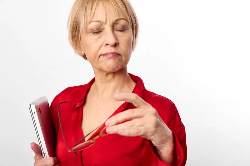 Elderly woman in red shirt holding glasses, gestures with raised hand, eyes closed in frustration or pain, isolated on white background, studio close up headshot serious