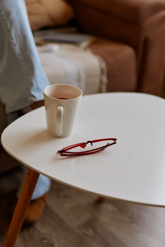 Casual woman at home sits in a cozy living room, round table holds a mug and glasses, soft natural light creates a calm, relaxed mood and everyday comfort. Subtle warmth adds depth to scene.