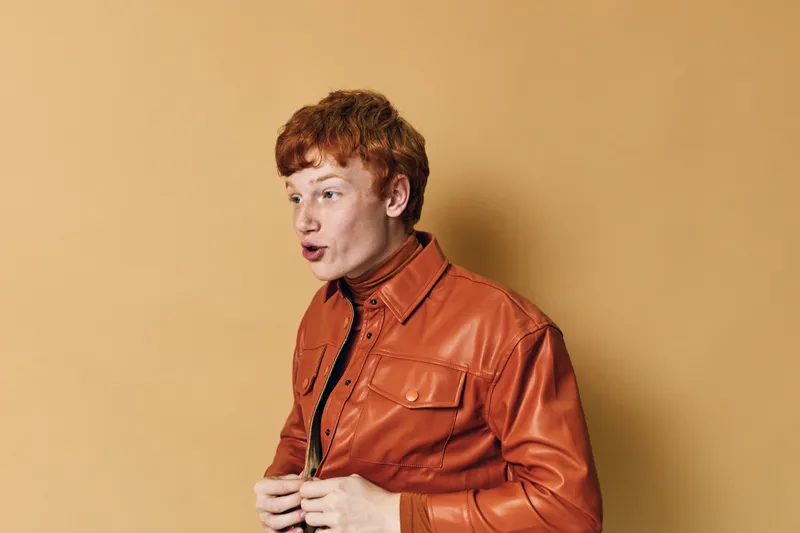 Young man with red hair wearing orange leather jacket posing and expressing surprise against warm yellow background in studio portrait photo.