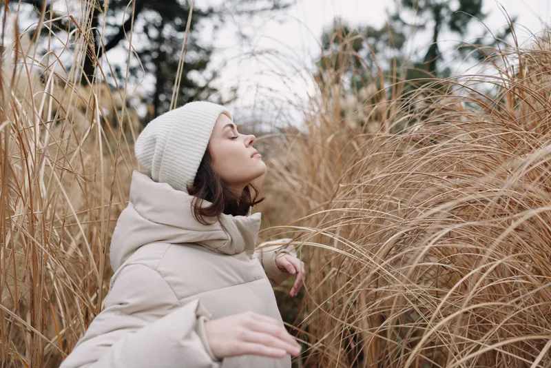 Woman in a warm coat and knit hat stands among tall golden grasses, embracing the quiet outdoor scene and brisk winter breeze under soft overcast light.