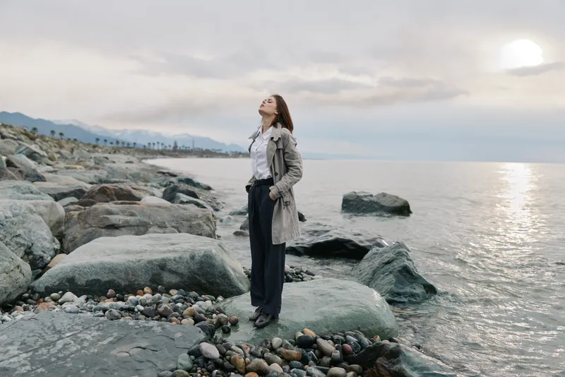 Young woman stands on rocky shore near calm sea under cloudy sky, wearing casual clothes. Concept of freedom, nature, and peaceful outdoor moment by the water landscape.