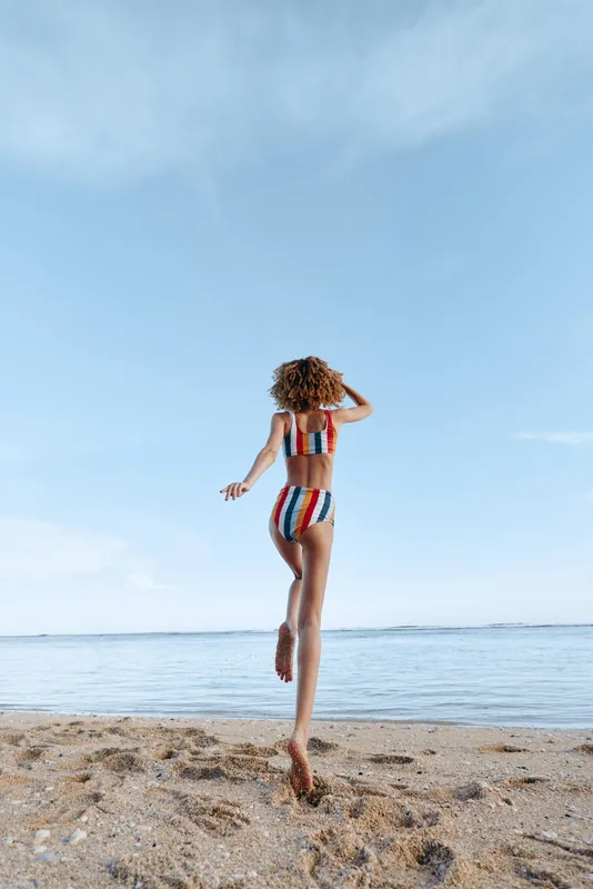 A young woman with curly hair runs barefoot on the sandy beach toward the calm ocean wearing a colorful striped swimsuit under a clear blue sky.