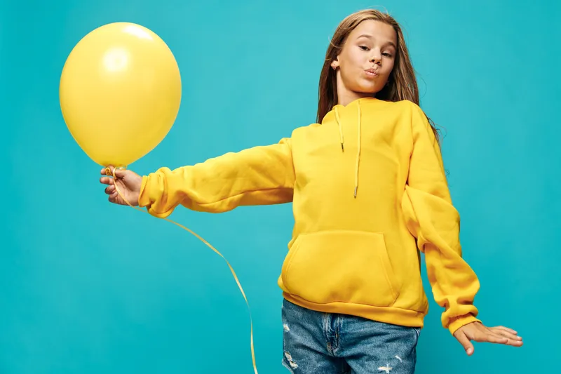 Girl with yellow balloon wearing yellow hoodie and jeans posing with closed eyes on blue background for celebration and fun. Child, birthday and happiness with casual clothes and balloon.