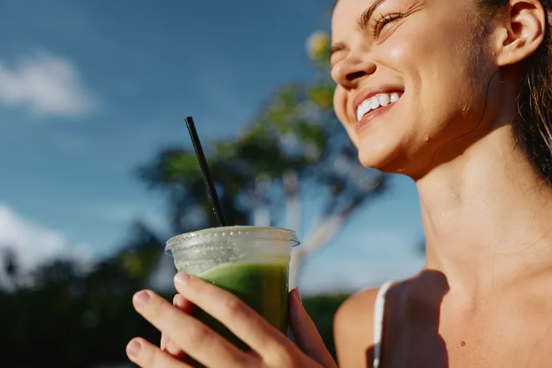 Happy woman enjoys a green smoothie outdoors, bright sun, casual summer vibe, holding a cup with straw, big smile, fresh flavors, lifestyle moment under clear blue sky.
