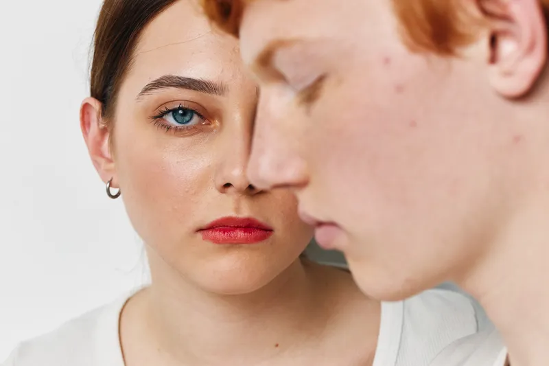 Woman with glowing skin and confident expression faces camera while young man with freckles looks down in closeup portrait on white background studio.