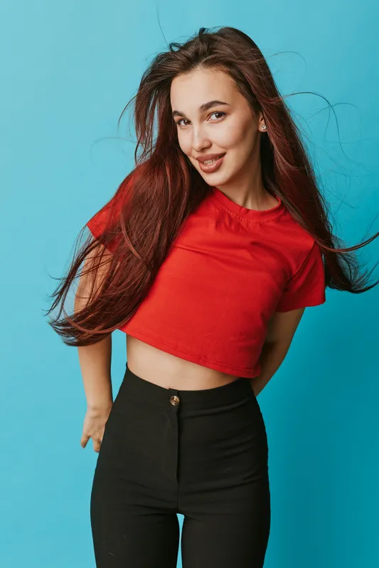 Young woman with long brown hair wearing red crop top and black pants smiling and posing against blue background in studio for fashion portrait and casual style.