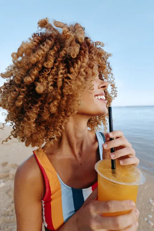 Curly haired woman at the beach smiles, holding iced coffee in a cup with straw, enjoying summer vacation and relaxation by the seaside, sunny outdoor lifestyle moment