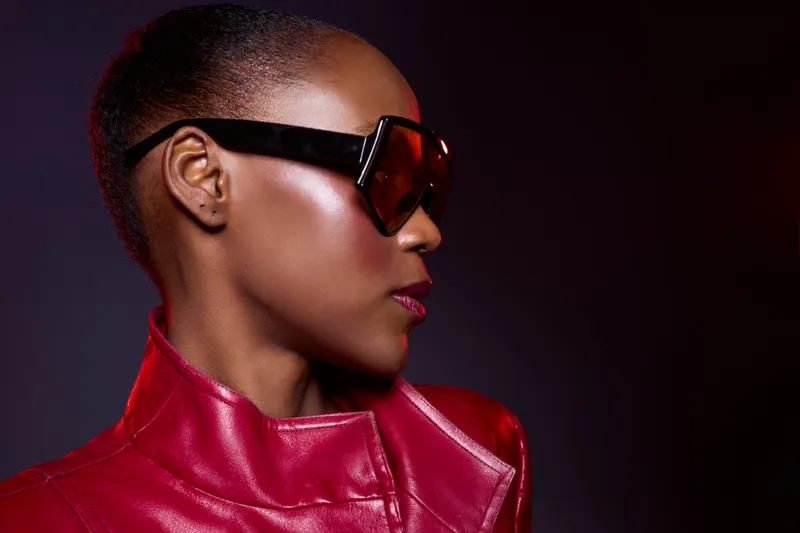 Profile of a dark skinned African woman wearing large black sunglasses and a shiny red leather jacket. Studio shot with dark background highlights fashion and modern style.