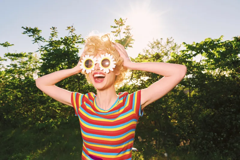 Smiling woman outdoors wearing a bright striped shirt and playful flower sunglasses basking in bright sunlight Retro color palette conveys cheerful lifestyle and carefree summer mood