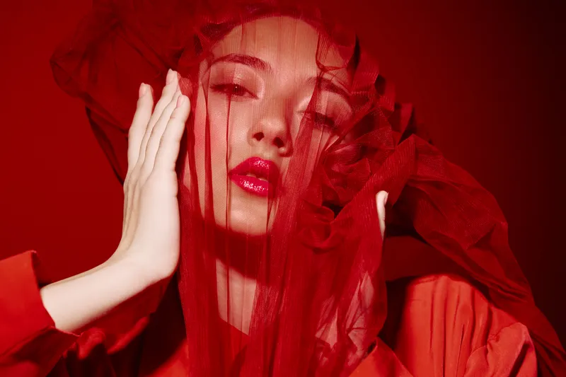 Fashion portrait of young woman with bright red tulle veil over face wearing red robe. Makeup emphasizes bold lips and sharp eyes. Concept of beauty, fashion, and creativity.