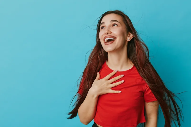 Young woman with long hair and red shirt smiling and looking up with hand on chest against blue background. Female model expressing happiness and positive emotion in studio.