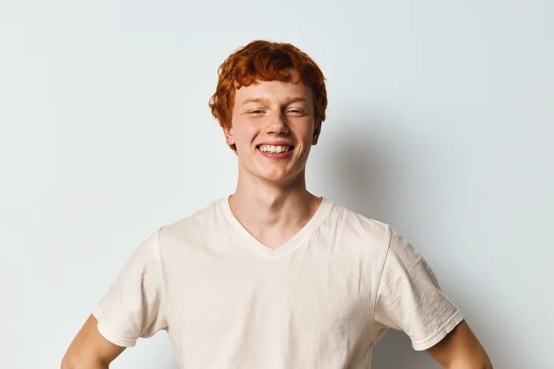Young man with red hair smiling wearing casual white t shirt standing against white background in studio portrait, happiness and positive emotion.