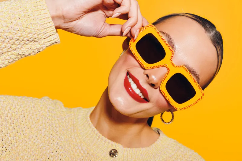 Smiling woman with bright red lipstick wears large yellow sunglasses and a beige sweater. Close-up portrait on vibrant yellow background. Concept of fashion, style, and summer mood.